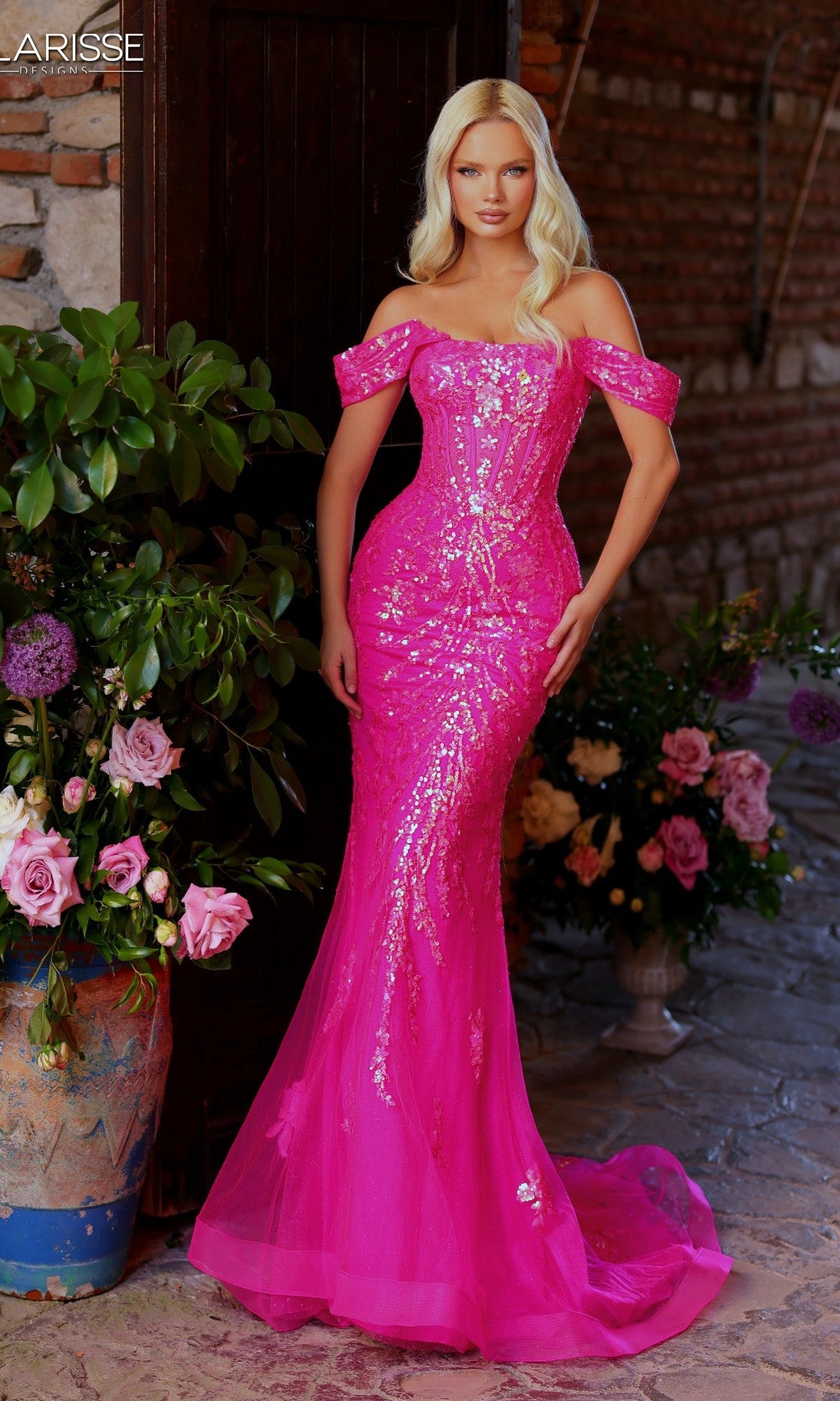 A woman in the Clarisse Designs 62245 long prom dress in iridescent fuchsia, featuring a mermaid silhouette, stands by a wooden door with potted flowers and brick walls behind her.